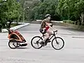 In China fährt dieser Vater mit einem Kind bei sich in der Tragetasche und einem Kind im Anhänger hinten dran.