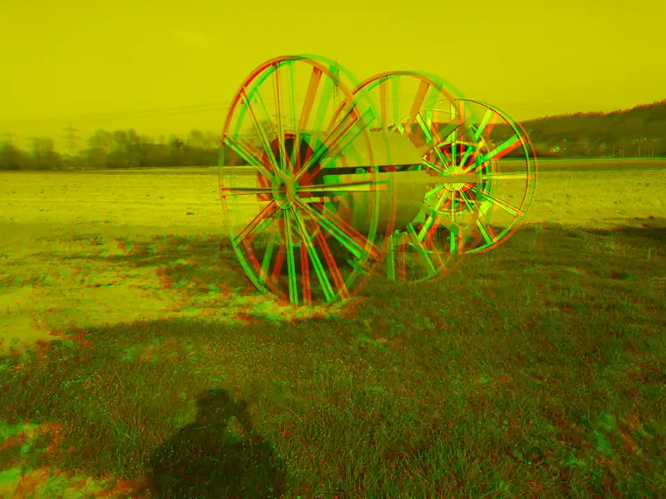 Anaglyph-3D-Bild von zwei Kabelrollen auf einem Feld. Dabei wurde der Hochspannungsmast über der vorderen Kabelrolle beiden Einzelbildern für das linke Auge und rechte Auge im Anaglyphbild zur Deckung gebracht.