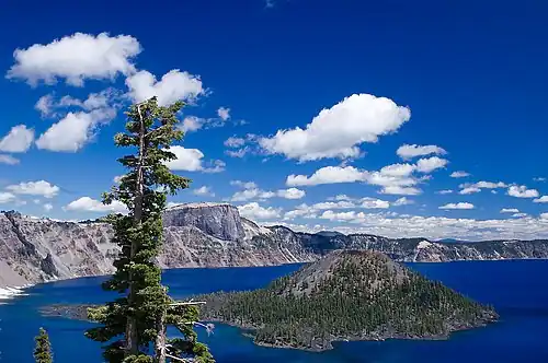 Wizard Island is shown in Crater Lake, with clouds in the sky above. The caldera rim appears to the left.